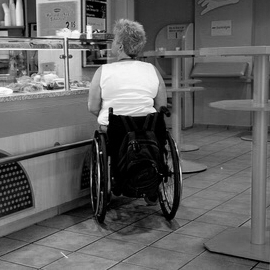Foto von einer Rollstuhlfahrerin an einer Imbisstheke ohne Unterfahrbarkeit, neben hohen Stehtischen